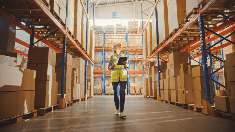 Professional Female Worker Wearing Hard Hat Checks Stock and Inventory with Digital Tablet Computer Walks in the Retail Warehouse full of Shelves with Goods